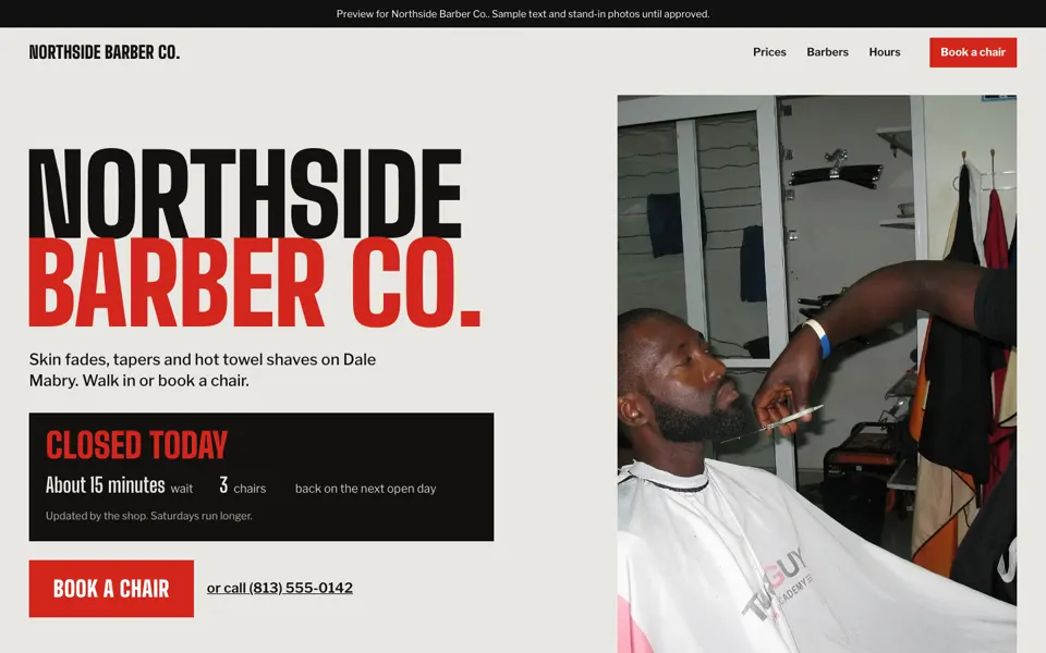 Barber shop homepage concept: a big red and black name, an open-or-closed wait board, and a Book a chair button beside a photo of a barber trimming a beard.